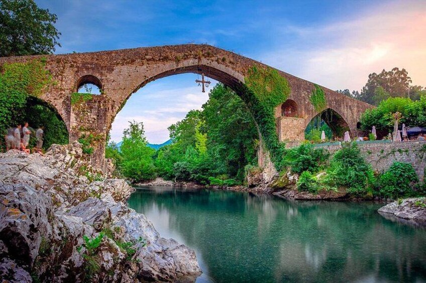 An ancient stone bridge with arches over a calm river, surrounded by lush greenery and rocky terrain. 2