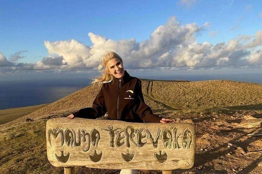 Una persona de pie detrás de un letrero de madera con texto, contemplando un paisaje montañoso con cielo despejado. 2