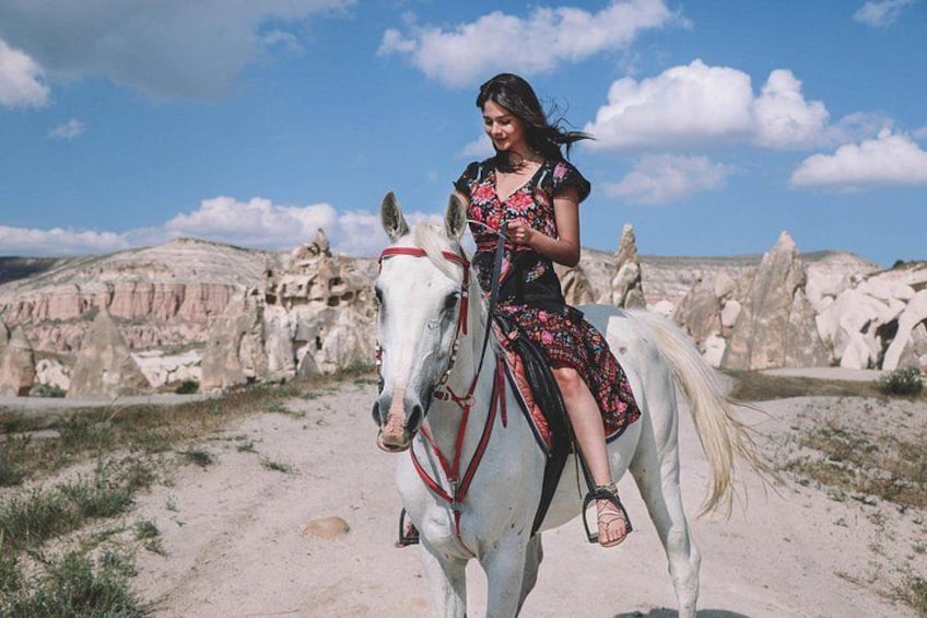Una persona montando un caballo blanco por un sendero de tierra, con formaciones rocosas al fondo. 4