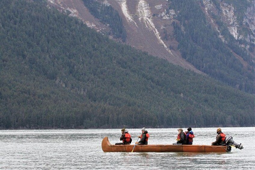 A longboat with four people rowing on a calm lake, surrounded by forested mountains. 4