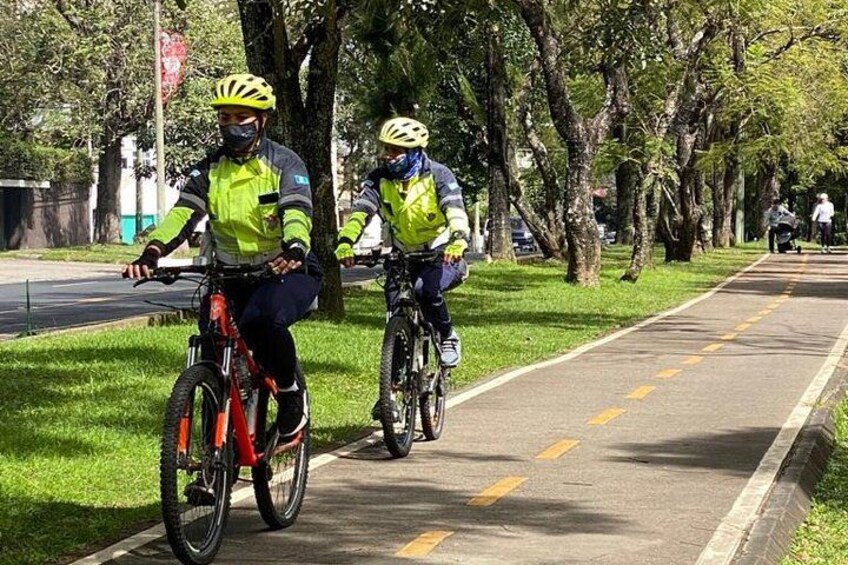 Two cyclists on a paved path with trees and grass on either side. 2