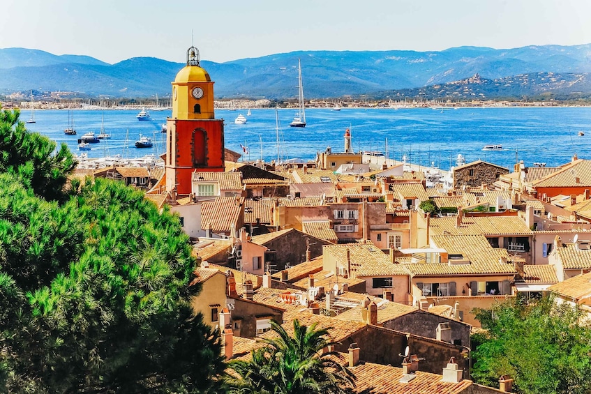 Una cittadina costiera con un'imponente torre dell'orologio gialla, edifici con tetti in terracotta e un porto turistico pieno di vele. 1