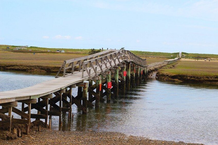 Un puente de madera sobre un río tranquilo, con orillas cubiertas de hierba y un cielo despejado. 2