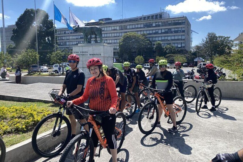 En grupp cyklister på ett offentligt torg, med ett monument och flaggor i bakgrunden. 5