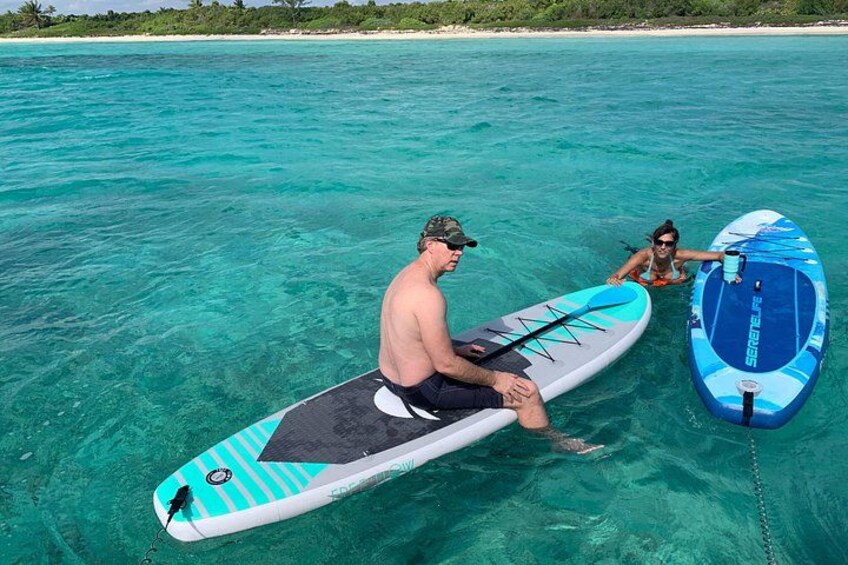Dos personas en tablas de remo en aguas turquesas cristalinas, con una playa y vegetación al fondo. 2
