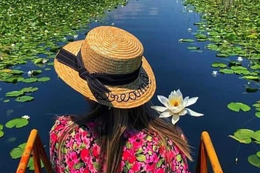 A person in a straw hat and floral dress, with a white flower in their hair, standing by a water body with lily pads. 1