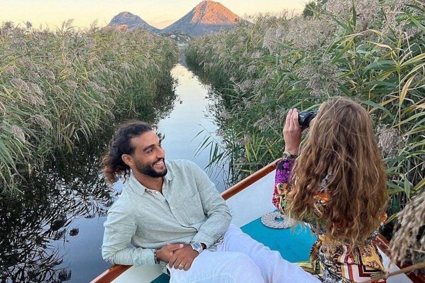 A river with dense vegetation on both sides, a boat with two people, and a mountainous landscape in the background. 1