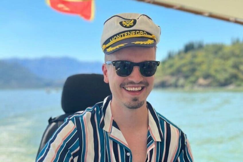 A person wearing a striped shirt and a cap with 'MONTENEGRO' on it, sitting on a boat with a body of water and a hilly landscape in the background. 5