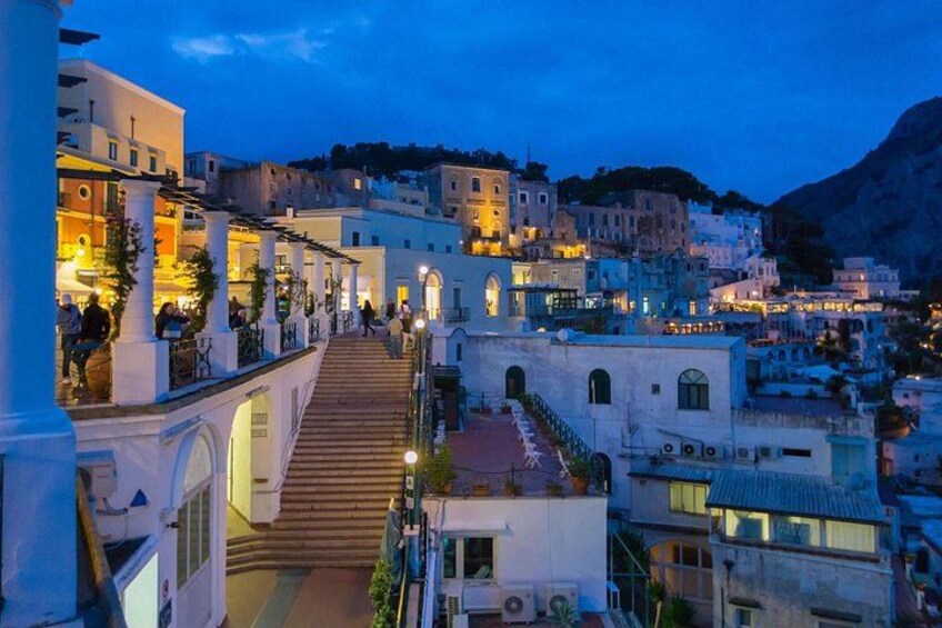 Un pueblo al atardecer con edificios iluminados, una escalera destacada y una montaña al fondo. 1