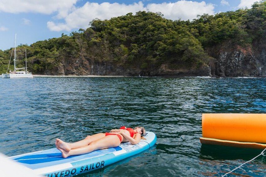 A person lying on a blue inflatable board labeled 'FIXEDO SAILOR' on calm water, with a boat and a rocky coastline in the background. 4