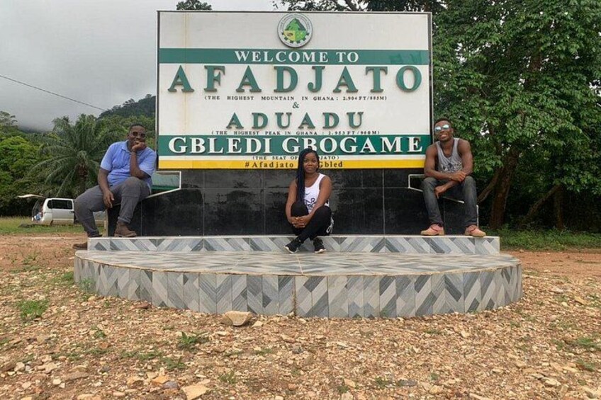 A welcome sign for Afadjato Aduadu Gbledi Grogame with three people sitting on a platform. 3