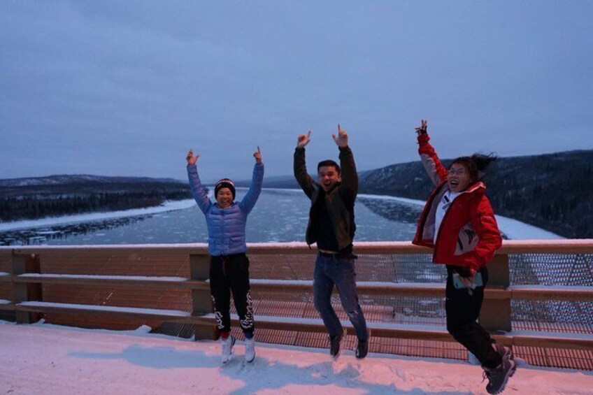 Tres personas saltando sobre un puente nevado, con un río helado y colinas al fondo. 2