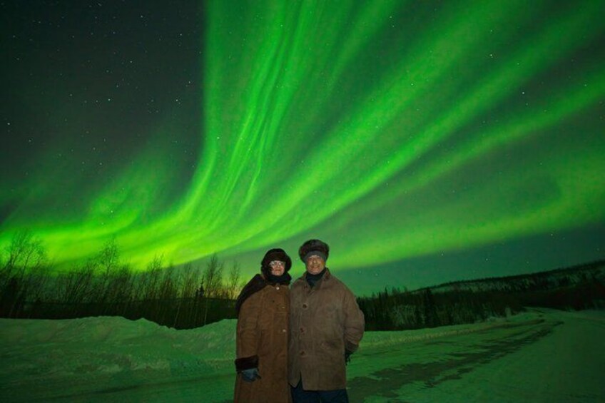 Two people standing on a snow-covered road under a green aurora display. 5