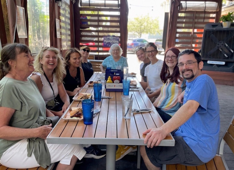 Un grupo de personas sentadas en una mesa de madera al aire libre, con bebidas y comida, en un patio cubierto con una parrilla al fondo. 3