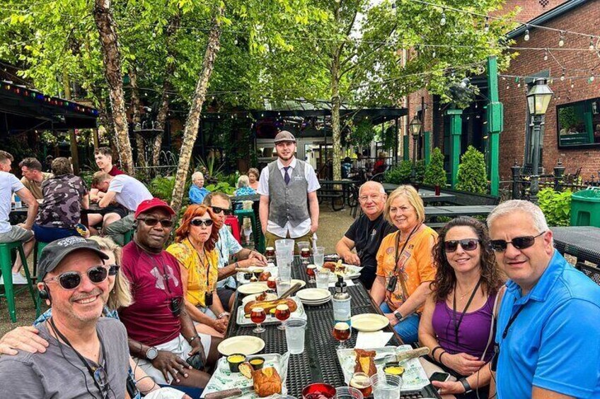 Un grupo de personas cenando al aire libre en un restaurante, con mesas y sillas, rodeado de árboles y edificios de ladrillo. 1