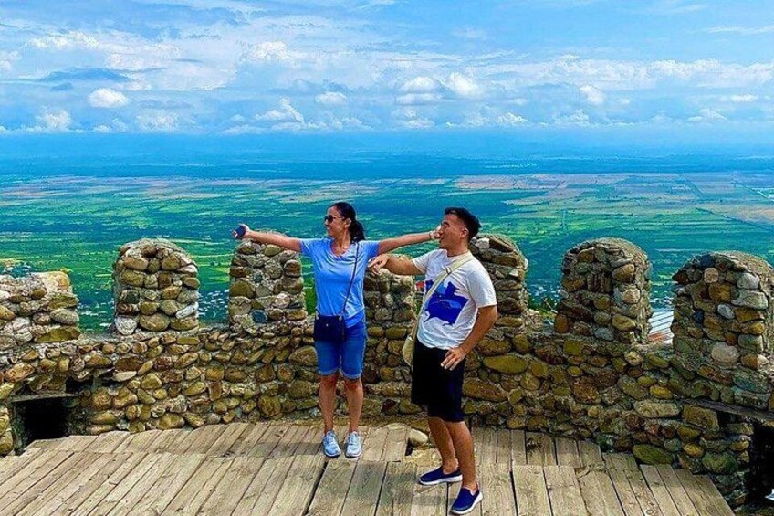 Due persone su una piattaforma di legno con un muro in pietra, che si affacciano su un paesaggio con campi e un orizzonte lontano. 1