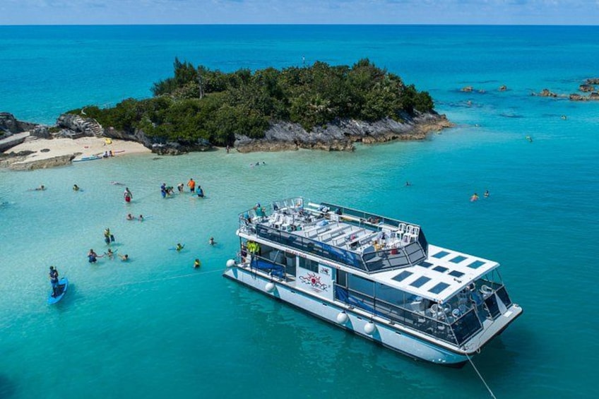 A large boat with people swimming and sunbathing in turquoise waters near a rocky island. 1