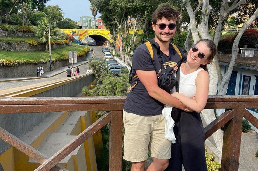 A couple standing on a bridge with a yellow pedestrian walkway, a road with cars, and a bridge with colorful decorations in the background. 2