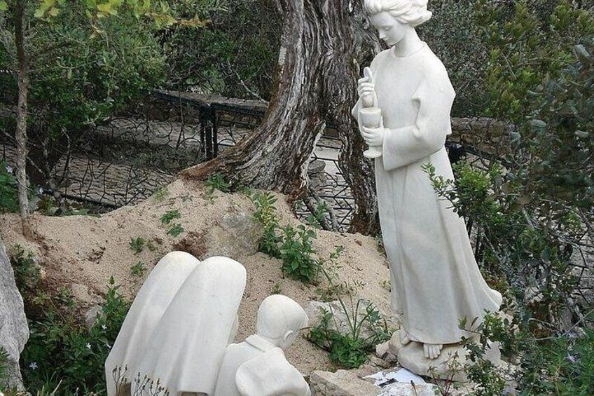 A white statue of a woman holding a book, surrounded by smaller statues and greenery. 2