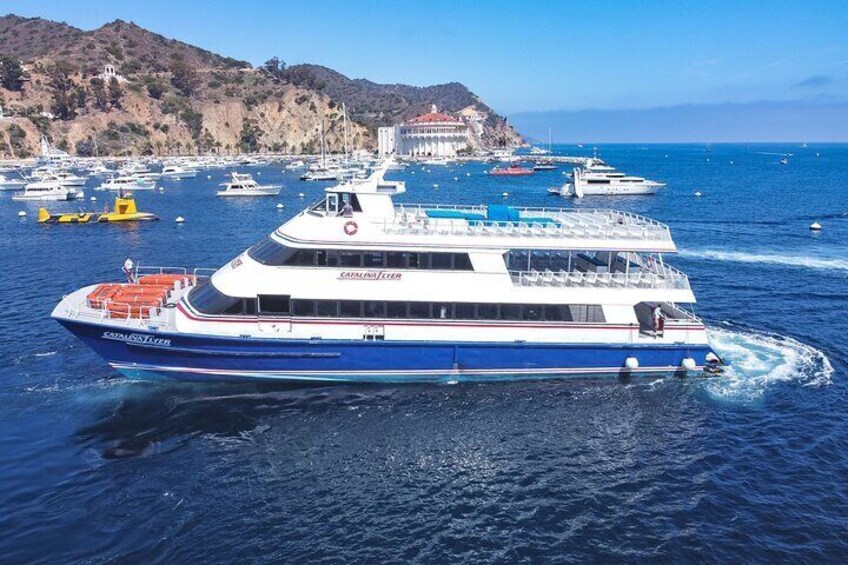 Un bote grande azul y blanco llamado 'Catalina Flyer' en el agua, con otros barcos y un pueblo costero al fondo. 5