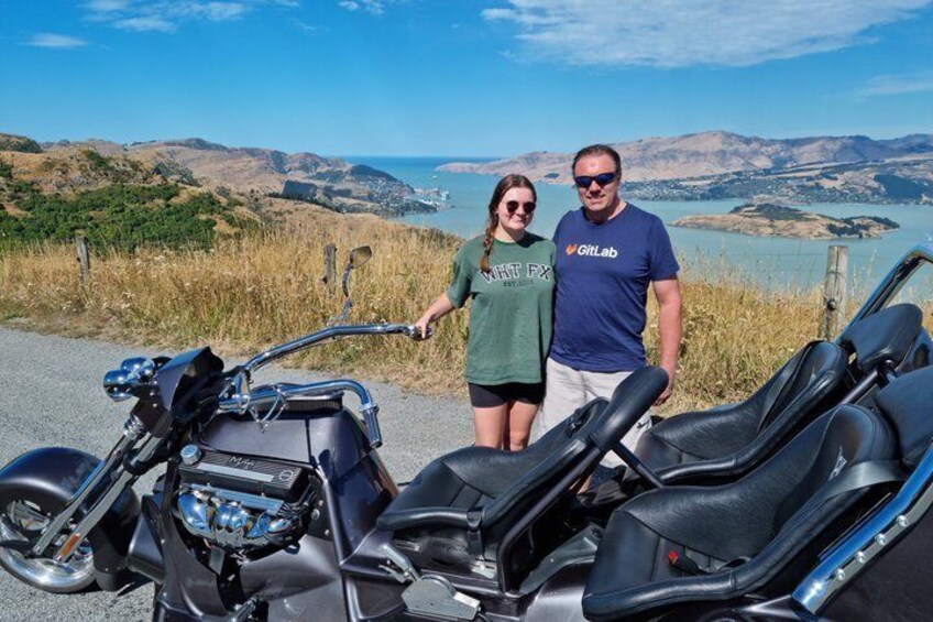 A motorcycle with a person standing beside it, a scenic view of hills and a body of water in the background. 1
