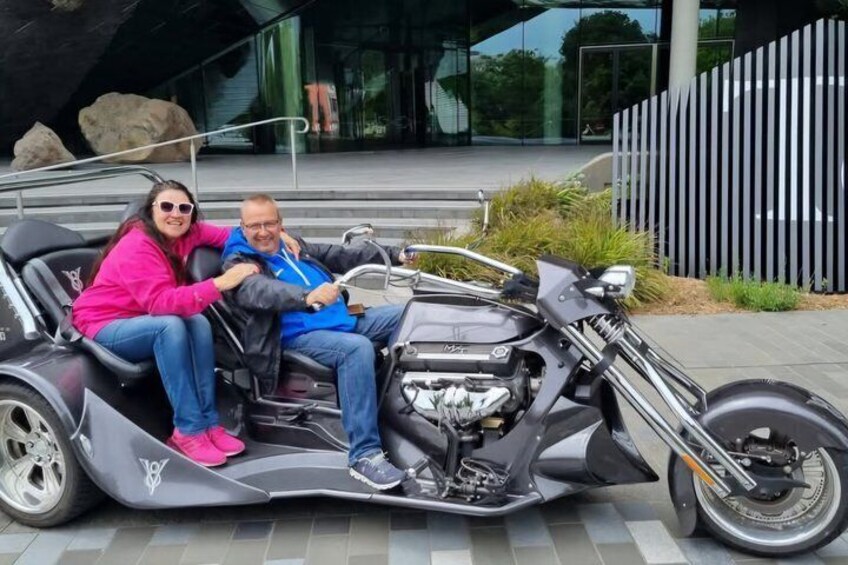 A man and a woman on a motorcycle with a sidecar, parked in front of a modern building with a glass facade. 5