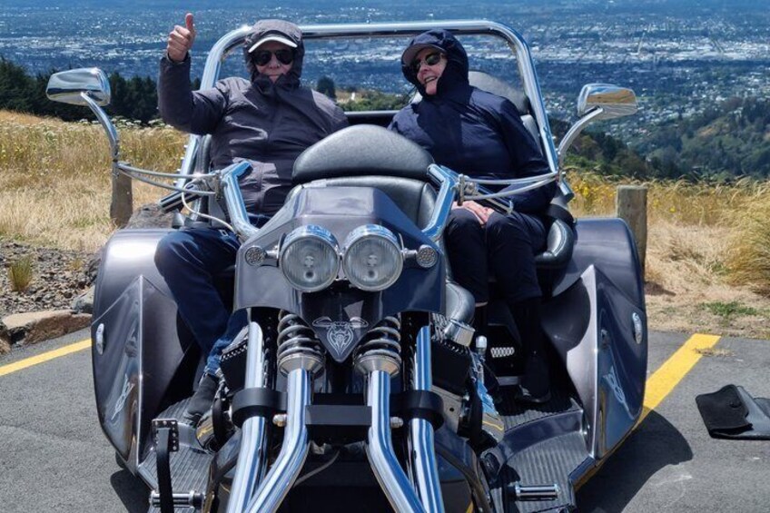 A vintage motorcycle with two riders, one giving a thumbs-up, on a road with a view of a city in the background. 2