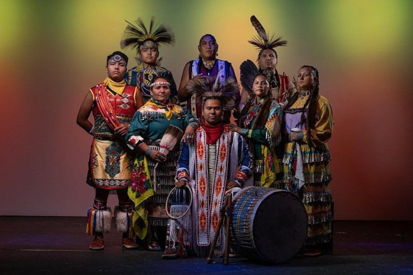 En grupp människor klädda i traditionella kläder från ursprungsbefolkningen i Nordamerika, några av dem håller i musikinstrument, på en scen med en färgstark bakgrund. 1