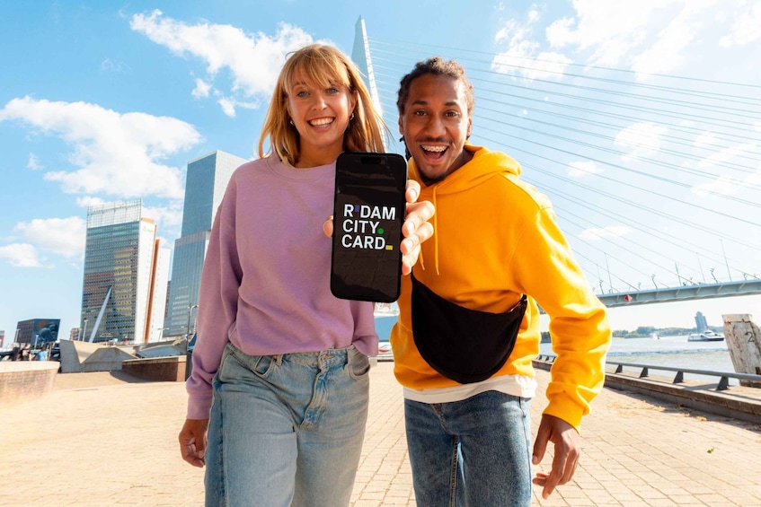 Two people holding a smartphone with 'RIDAM CITY CARD' on the screen, standing in front of a modern bridge and city buildings. 1