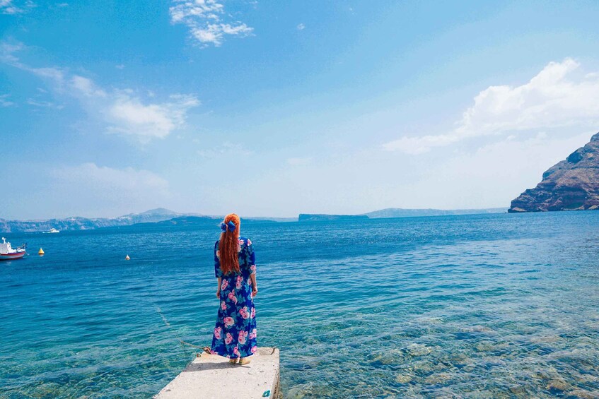 A person in a blue floral dress standing on a pier, overlooking a calm sea with boats and distant mountains. 5