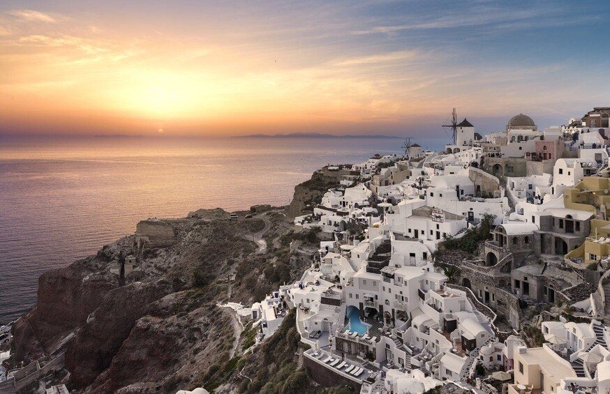 A coastal village with white buildings, a cliffside, and the sea at sunset. 1