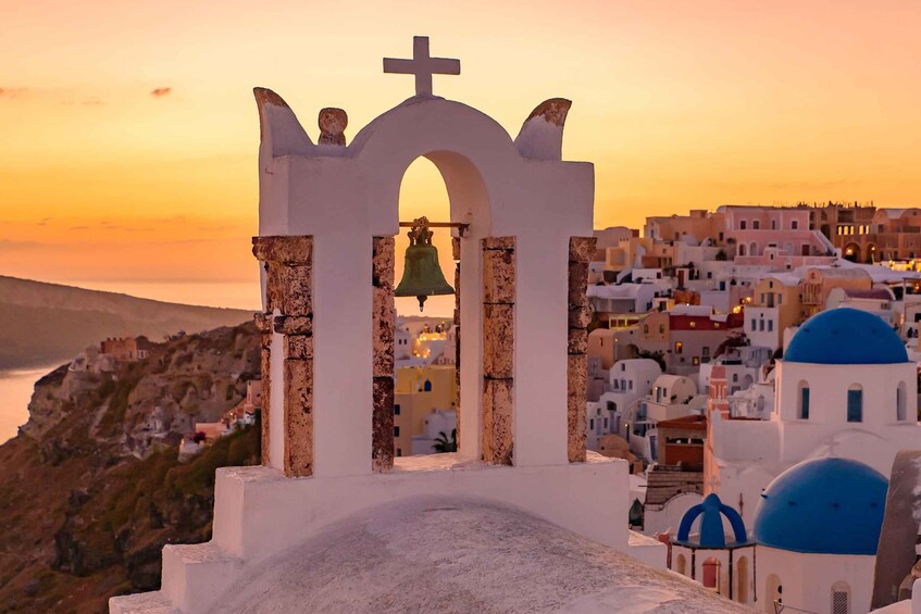 A white church with a bell tower, blue domes, and a view of a coastal town at sunset. 2