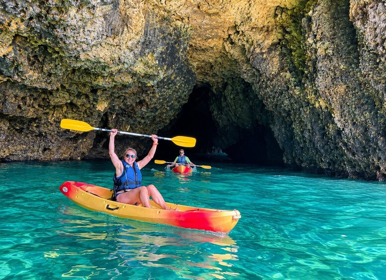 Two people kayaking in clear turquoise water with a rocky cave in the background. 3