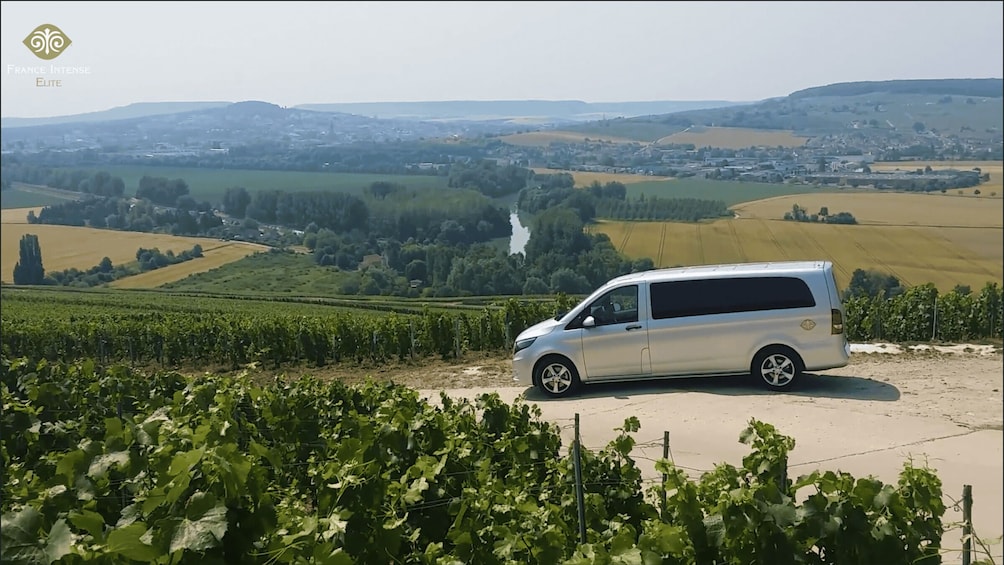 A silver minivan parked on a dirt road overlooking vineyards and a rural landscape. 2