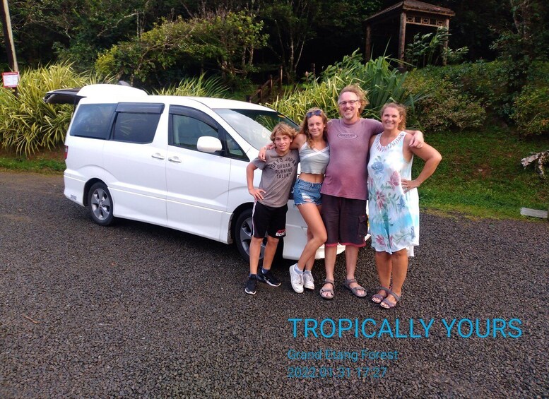 Una minivan blanca estacionada en un camino de grava, con un grupo de personas de pie junto a ella. El fondo muestra un follaje exuberante y una estructura de madera. El texto de la imagen dice: TROPICALLY YOURS Grand Etang Forest 2022 01 31 17:27. 5