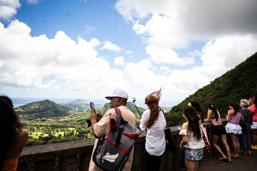 Un groupe de personnes prend des photos sur un belvédère offrant une vue panoramique sur une vallée verdoyante et des montagnes lointaines. 2