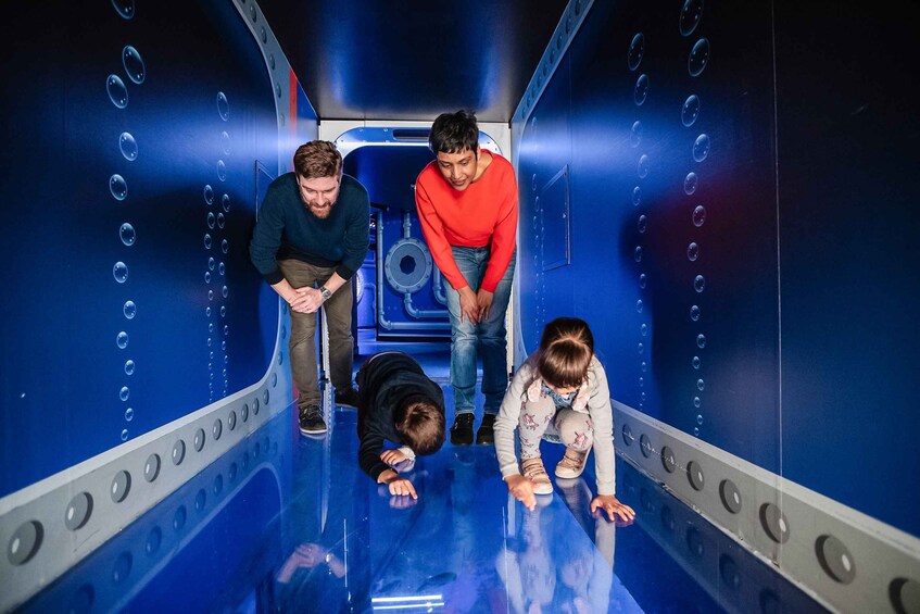 Four people in a blue, cylindrical, metallic structure with circular patterns on the walls and floor. 3