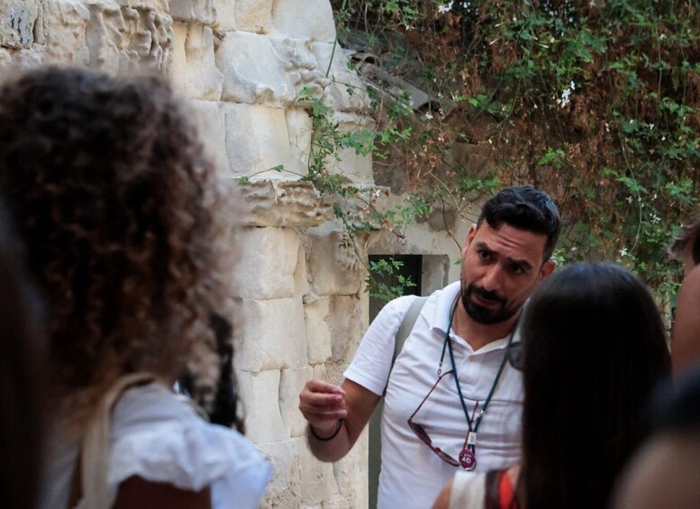 A man in a white shirt and glasses standing in front of a stone wall with greenery growing on it. 5