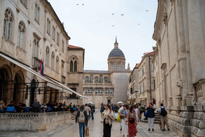 A historic street with people walking, a prominent building with a dome, and a café with patrons. 1