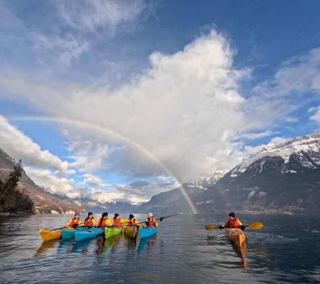 Een groep kajakkers in kleurrijke kajaks op een kalm meer, met een regenboog op de achtergrond en besneeuwde bergen. 4