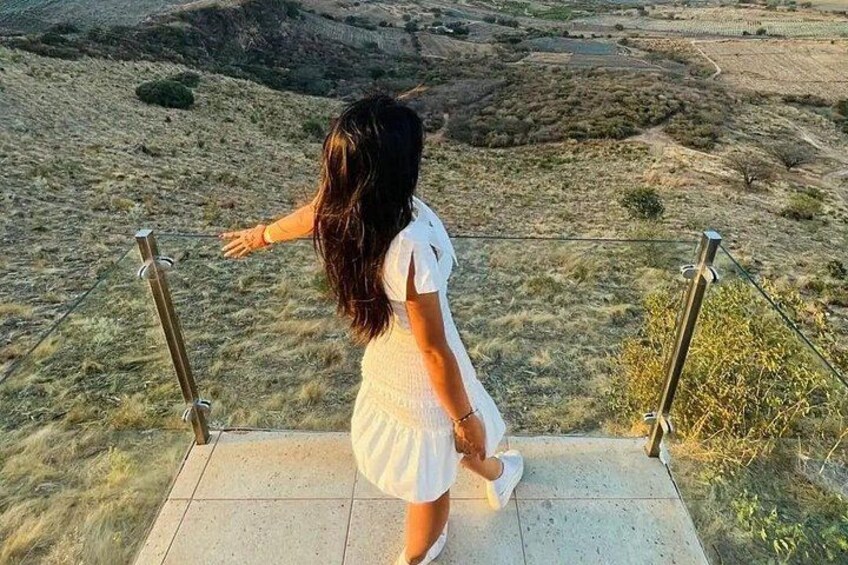 A person in a white dress standing on a concrete path with metal railings, overlooking a hilly landscape with sparse vegetation. 4