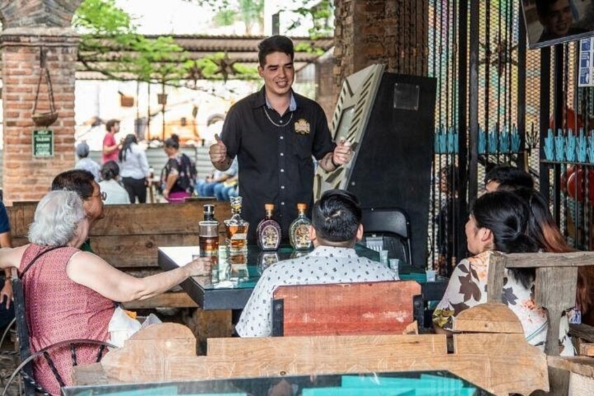 A person standing behind a table with bottles, a wooden bench, and a brick wall. 2