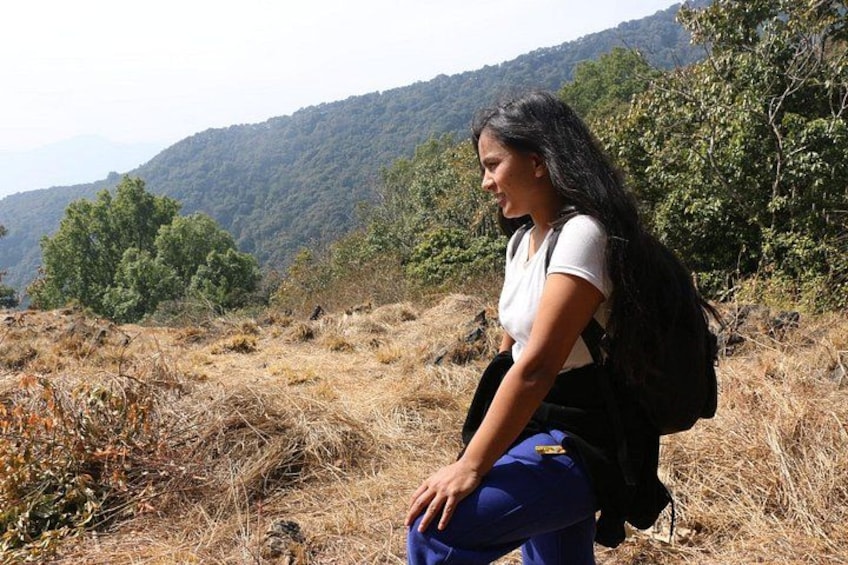 A person crouching on a hillside with a backpack, surrounded by dry grass and trees. 5