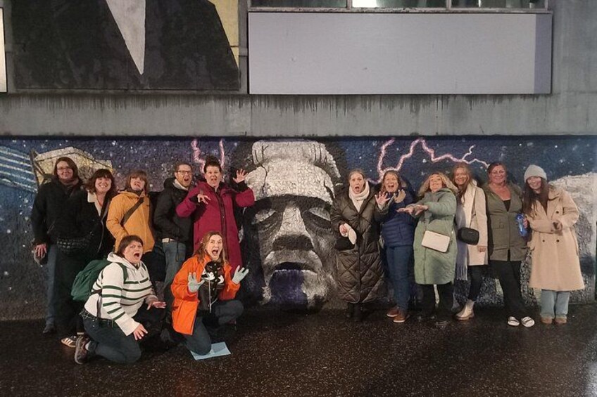 A group of people posing in front of a mural with a large face and lightning. 1