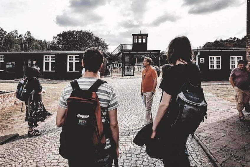 A group of people walking on a cobblestone path, a building with a tower, and a fence in the background. 5
