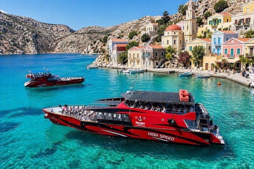 Ein rotes Boot namens 'Rhodes' mit dem Aufschrift 'High Speed', liegt im Hafen vor türkisfarbenem Wasser und bunten Gebäuden. 1