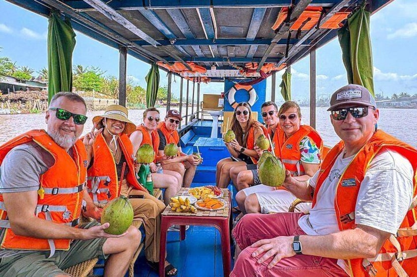 Eine Gruppe von Menschen auf einem Boot, mit Rettungswesten, wobei auf dem Tisch Kokosnüsse und Früchte liegen. 2