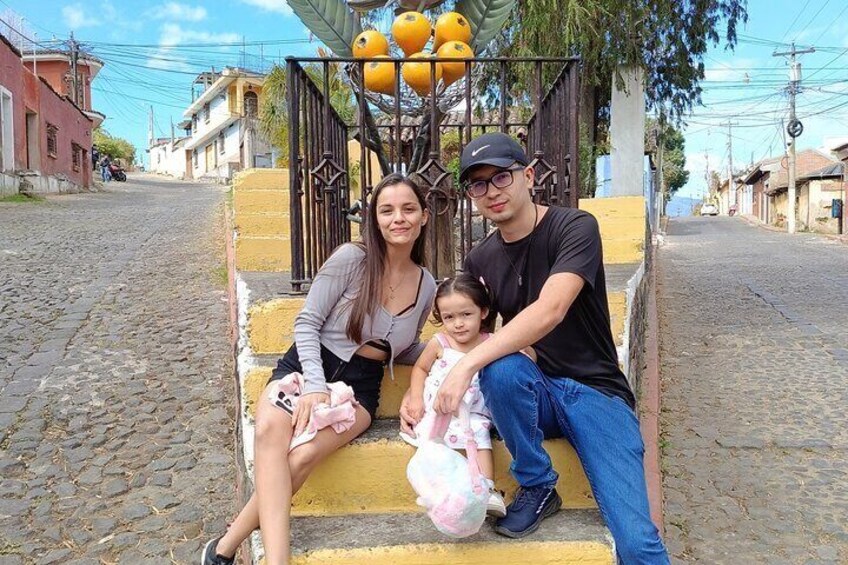 A family of three on a cobblestone street with buildings and a gate with oranges in the background. 4