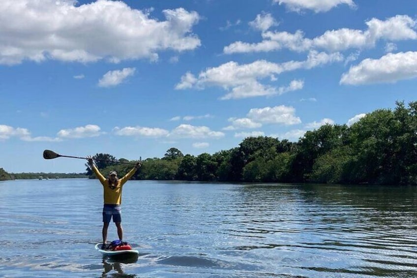 Una persona in piedi su una tavola da paddleboard in acqua, con un remo in mano, sullo sfondo alberi e un cielo sereno. 2