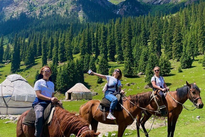 Three people riding horses in a mountainous area with tents and pine trees in the background. 1